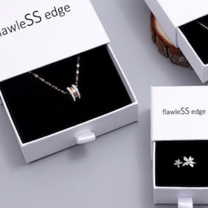 Peut inclure: Deux bo&icirc;tes blanches avec des int&eacute;rieurs noirs et le texte "flawlessS edge" sur le devant. La bo&icirc;te du haut est ouverte et contient un collier en argent avec un pendentif rond. La bo&icirc;te du bas est ferm&eacute;e et contient une paire de petites boucles d'oreilles en argent.