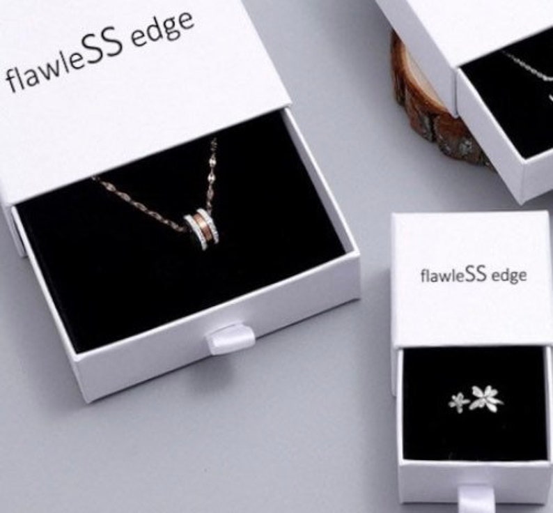 Peut inclure: Deux bo&icirc;tes blanches avec des int&eacute;rieurs noirs et le texte "flawlessS edge" sur le devant. La bo&icirc;te du haut est ouverte et contient un collier en argent avec un pendentif rond. La bo&icirc;te du bas est ferm&eacute;e et contient une paire de petites boucles d'oreilles en argent.