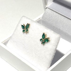 May include: A pair of gold butterfly earrings with green emerald stones. The earrings are in a white jewelry box.