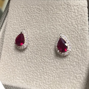 Può includere: Un paio di orecchini d'argento con una pietra preziosa di rubino a forma di lacrima circondata da un alone di pietre preziose più piccole e trasparenti.