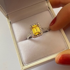 May include: A silver ring featuring a large, yellow, emerald-cut gemstone surrounded by a halo of smaller diamonds.