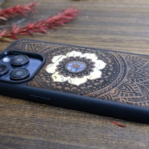 以下が含まれることがあります： 黒のゴム製バンパーが付いた茶色の木製携帯電話ケース。ケースには、中央に白い花が咲いた彫刻されたマンダラ模様が施されています。花には青と緑のアワビの貝殻の象嵌が施されています。