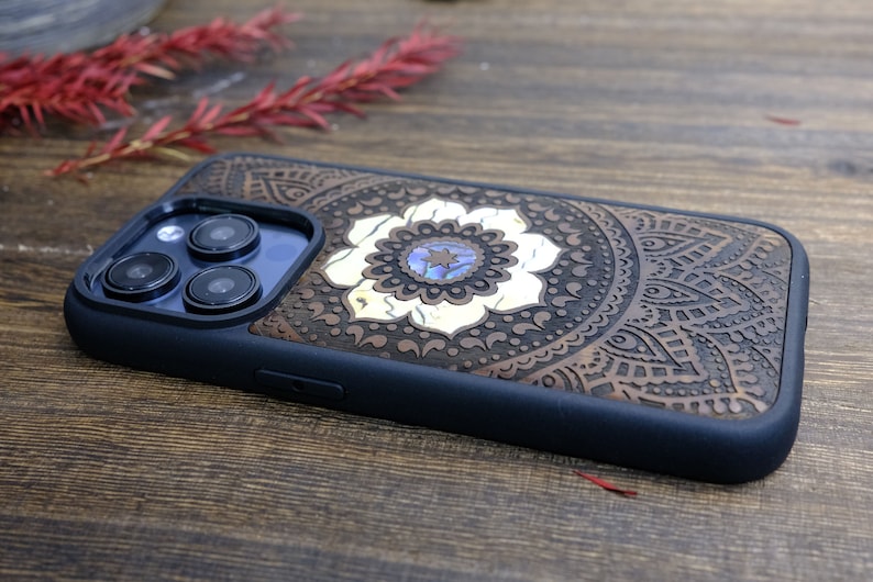 以下が含まれることがあります： 黒のゴム製バンパーが付いた茶色の木製携帯電話ケース。ケースには、中央に白い花が咲いた彫刻されたマンダラ模様が施されています。花には青と緑のアワビの貝殻の象嵌が施されています。