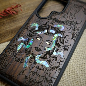Op de afbeelding: Een donkerhouten telefoonhoes met een zwarte rand. De hoes heeft een gedetailleerde gravure van Medusa met slangen en bloemen. De slangen zijn ingelegd met iriserende parelmoer.