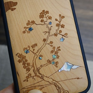 K&ouml;nnte beinhalten: Eine Handyh&uuml;lle aus Holz mit einem Laser-geschnittenen Design eines Baumes mit Blumen und einem Berg. Die Blumen sind mit irisierendem Perlmutt eingelegt.
