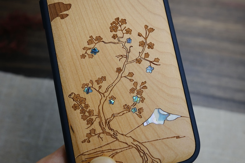 K&ouml;nnte beinhalten: Eine Handyh&uuml;lle aus Holz mit einem Laser-geschnittenen Design eines Baumes mit Blumen und einem Berg. Die Blumen sind mit irisierendem Perlmutt eingelegt.