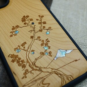 K&ouml;nnte beinhalten: Eine Handyh&uuml;lle aus Holz mit schwarzem Gummibumper. Die H&uuml;lle zeigt ein lasergraviertes Design eines Baumes mit Blumen und einer Bergszene. Die Blumen sind mit irisierendem Perlmutt eingelegt.