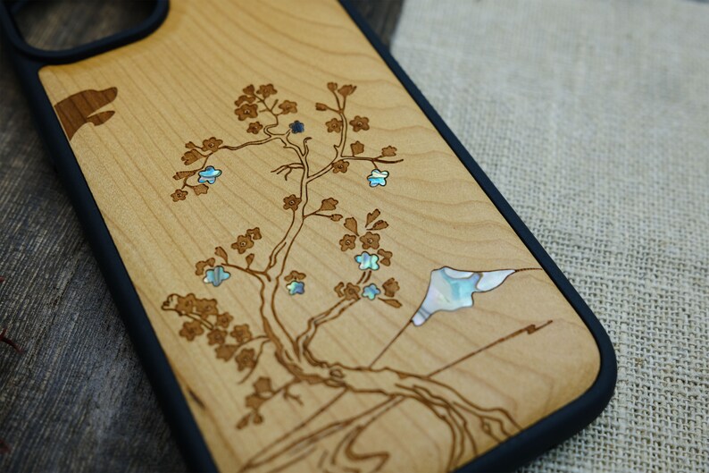 K&ouml;nnte beinhalten: Eine Handyh&uuml;lle aus Holz mit schwarzem Gummibumper. Die H&uuml;lle zeigt ein lasergraviertes Design eines Baumes mit Blumen und einer Bergszene. Die Blumen sind mit irisierendem Perlmutt eingelegt.