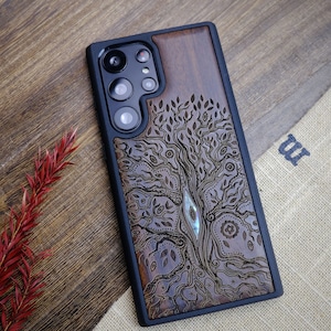 May include: A dark brown wooden phone case with a carved tree design. The case has a black border and a cutout for the phone's camera.