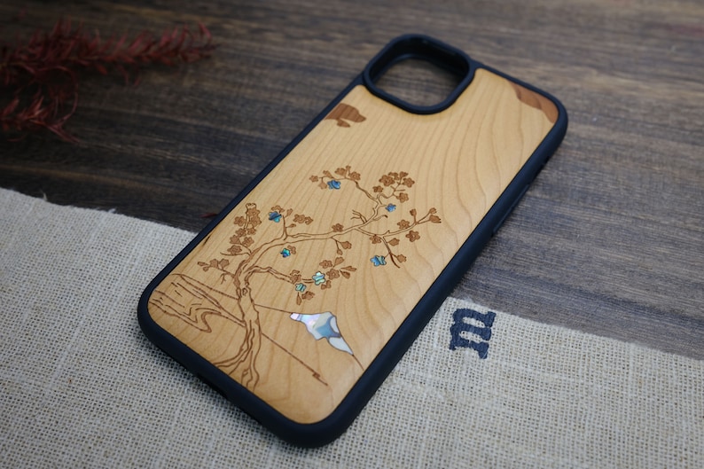 K&ouml;nnte beinhalten: Eine Handyh&uuml;lle aus Holz mit schwarzem Gummibumper. Die H&uuml;lle zeigt ein geschnitztes Design eines Baumes mit blauen und gr&uuml;nen Akzenten.
