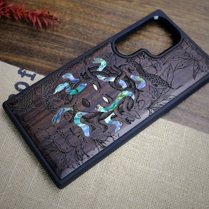 Op de afbeelding: Een donkerhouten telefoonhoes met een zwarte rubberen bumper. De hoes heeft een gedetailleerde gravure van Medusa met slangen en bloemen, geaccentueerd met iriserende parelmoer.