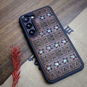 May include: A brown wooden phone case with a black border and a geometric pattern. The case is decorated with small, iridescent seashells in the shape of turtles.