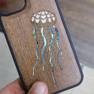 May include: A wooden phone case with a black border and a detailed, iridescent abalone inlay of a jellyfish.