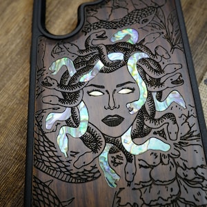 Op de afbeelding: Een houten telefoonhoesje met een zwarte rand en een gedetailleerde gravure van Medusa met slangen in haar haar. De slangen zijn gemaakt van iriserende abalone-parelmoer.