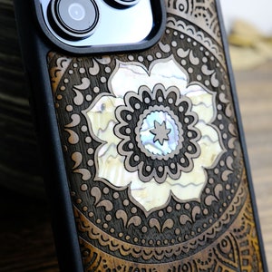 Pode incluir: Uma capa de telefone preta com um design de madeira marrom e uma incrustação de flor branca. A flor tem um centro azul e branco.