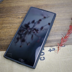 Op de afbeelding: Een zwarte smartphone met een schermbeschermer op een jute oppervlak. De telefoon reflecteert een wazig beeld van een rode plant.