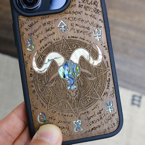 May include: A wooden phone case with a black border and a detailed design of a goat's head with horns and a pentagram. The design is made of wood and mother of pearl.