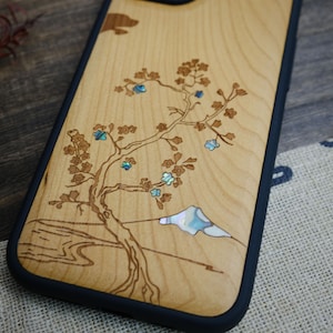 K&ouml;nnte beinhalten: Eine Handyh&uuml;lle aus Holz mit einem lasergravierten Design eines Baumes mit blauen Bl&uuml;ten und einem Berg. Das Design ist mit Perlmutt eingelegt.