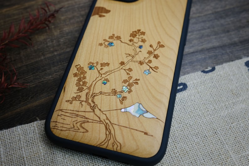 K&ouml;nnte beinhalten: Eine Handyh&uuml;lle aus Holz mit einem lasergravierten Design eines Baumes mit blauen Bl&uuml;ten und einem Berg. Das Design ist mit Perlmutt eingelegt.