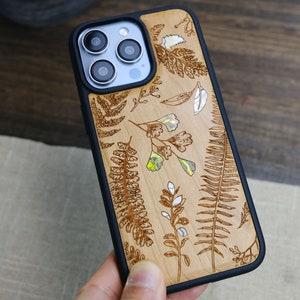 May include: A wood phone case with a black border and a floral design. The design features ferns and other plants in a light brown color.