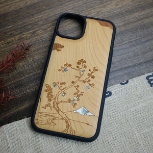 K&ouml;nnte beinhalten: Eine Handyh&uuml;lle aus Holz mit schwarzem Gummibumper. Die H&uuml;lle zeigt ein lasergraviertes Design eines Baumes mit einem Berg im Hintergrund. Der Baum und der Berg sind mit Perlmutt eingelegt.