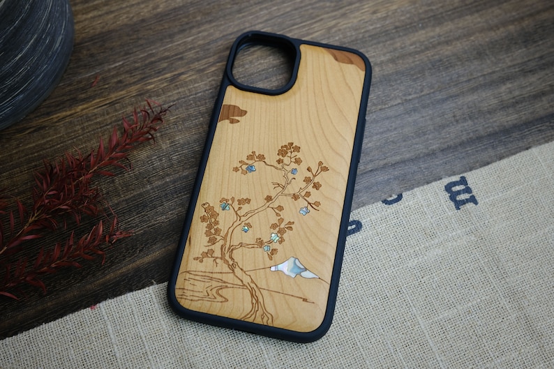 K&ouml;nnte beinhalten: Eine Handyh&uuml;lle aus Holz mit schwarzem Gummibumper. Die H&uuml;lle zeigt ein lasergraviertes Design eines Baumes mit einem Berg im Hintergrund. Der Baum und der Berg sind mit Perlmutt eingelegt.