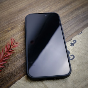 K&ouml;nnte beinhalten: Ein schwarzes Smartphone mit einem Displayschutz auf einer Holzoberfl&auml;che. Das Telefon befindet sich in einem schwarzen Etui.