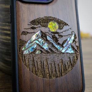 Può includere: Una custodia per telefono in legno con un design circolare che presenta una scena di montagna con una luna e nuvole. Le montagne sono intarsiate con madreperla di abalone iridescente.