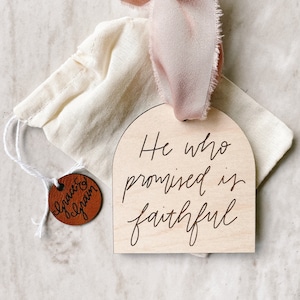 Puede incluir: Un letrero de madera con la parte superior redondeada y el texto "He who promised is faithful" escrito en cursiva. El letrero está atado con una cinta rosa y viene con una pequeña etiqueta de cuero marrón que dice "grace &  grain".