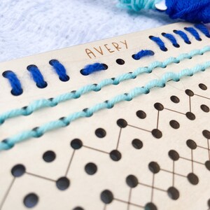 以下が含まれることがあります： 糸を織るための穴のグリッドが付いた木製のボード。ボードには「AVERY」という名前が書かれています。青い糸とターコイズブルーの糸が穴に通されています。