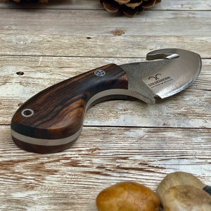 May include: A knife with a brown wooden handle and a curved blade with a hook at the end. The blade is made of stainless steel and has the words "GOLDENHORN" engraved on it. The knife is resting on a wooden surface.