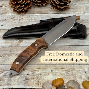Può includere: Un coltello da caccia artigianale con manico in legno e lama in acciaio inossidabile. Il coltello è appoggiato su una superficie in legno con pigne e pietre.