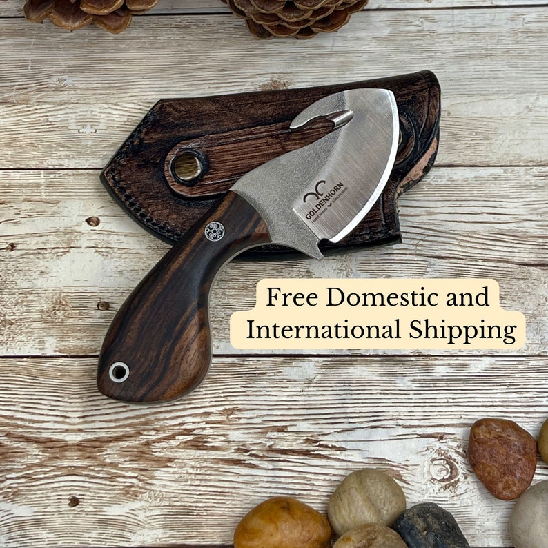 May include: A small, curved knife with a wooden handle and a leather sheath. The blade has a hook on the end and the handle is made of dark wood with a light wood inlay. The knife is engraved with the words "Goldenhorn Handmade in Canada".