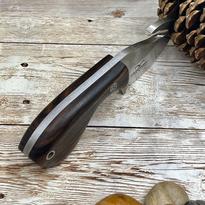 May include: A close-up of a silver knife with a dark wood handle. The knife has a small hook on the end of the blade and the text "COLD STEEL" is engraved on the blade.