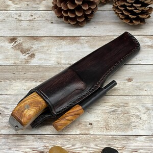 Pu&ograve; includere: Un fodero in pelle marrone con un coltello con manico in legno e un accendino nero all'interno. Il fodero &egrave; decorato con cuciture e l'accendino ha un manico in legno.