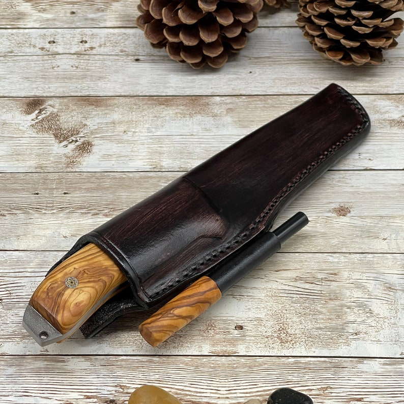 Pu&ograve; includere: Un fodero in pelle marrone con un coltello con manico in legno e un accendino nero all'interno. Il fodero &egrave; decorato con cuciture e l'accendino ha un manico in legno.