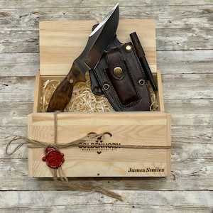 May include: A wooden box with a brown leather sheath and a knife inside. The box is engraved with the text "Goldenhorn expert in handcraft" and "James Smiles".