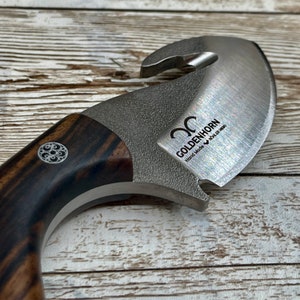 May include: A close-up of a brown and black wooden handled knife with a stainless steel blade. The blade has a unique shape with a small hook on the top and a curved edge. The knife is engraved with the words "Goldenhorn Hand Made B&ouml;hler N690".