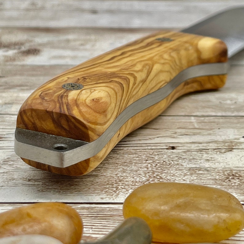 Pu&ograve; includere: Primo piano di un manico di coltello in legno con una fascia di metallo argentato. Il manico &egrave; in legno di ulivo con una venatura naturale.