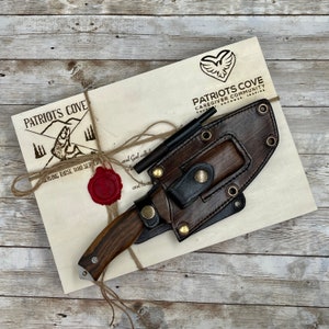 May include: A brown leather sheath with a knife and a fire starter inside. The sheath has a brass button and stitching. The knife has a wooden handle and a metal blade. The fire starter is a small metal rod. The sheath is on a wooden surface with a piece of paper that says "Patriots Cove Caregiver Community" and "Serving those who served".