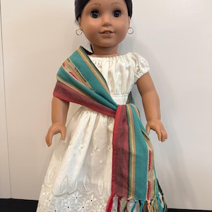 May include: A doll with dark brown hair and brown eyes wearing a white off-the-shoulder dress with an embroidered pattern and a scalloped hem. A woven sash featuring teal, maroon, and tan stripes with fringed ends is draped across the torso.