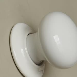 Op de afbeelding: Een close-up van een witte keramische deurknop. De knop is rond en glad, met een gebogen basis die aan de deur bevestigd is. De knop is gemonteerd op een lichtbeige oppervlak.