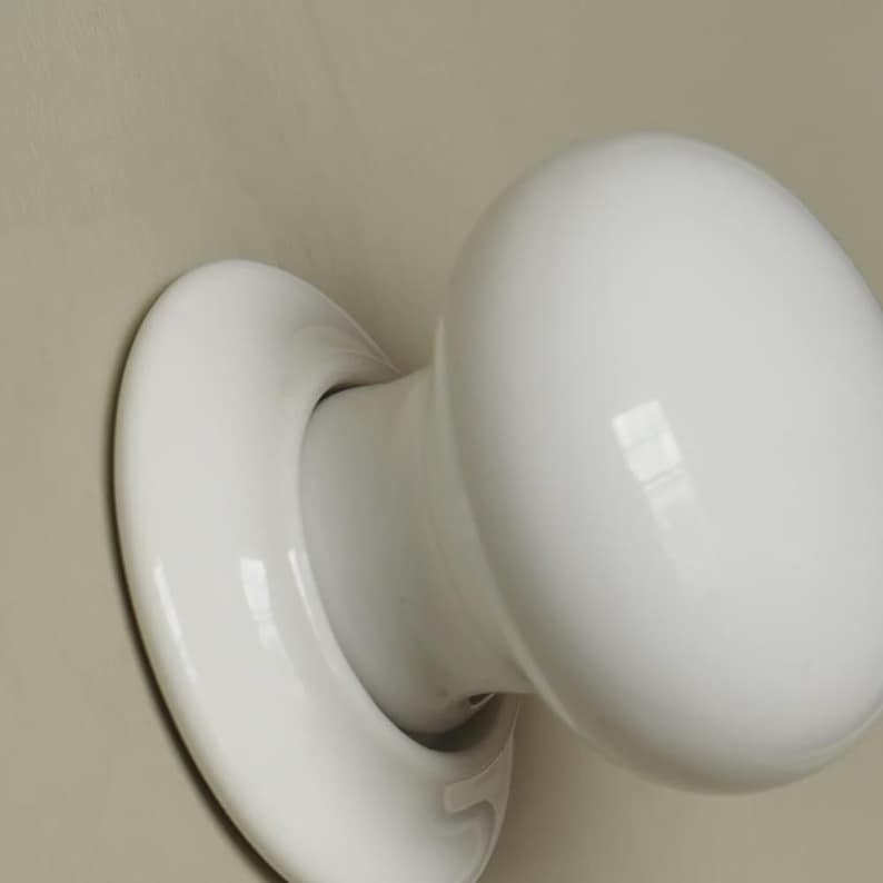 Op de afbeelding: Een close-up van een witte keramische deurknop. De knop is rond en glad, met een gebogen basis die aan de deur bevestigd is. De knop is gemonteerd op een lichtbeige oppervlak.