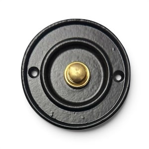 Può includere: Un campanello circolare con una finitura nera opaca sulla piastra esterna e un pulsante centrale color ottone. La piastra presenta due piccoli fori per le viti sui lati opposti e un design a cerchi concentrici in rilievo attorno al pulsante.