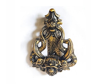 Könnte beinhalten: Ein goldener und schwarzer Türklopfer aus Metall mit einem detaillierten Design, das eine mythische Kreatur mit Hörnern und Bart zeigt. Der Türklopfer hat die Form eines stilisierten Gesichts mit komplizierten Details.
