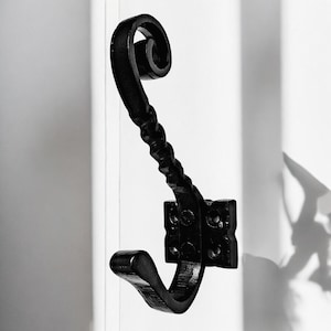 Pode incluir: Gancho de metal preto com um design decorativo de volutas. O gancho é montado em uma parede com uma base quadrada.