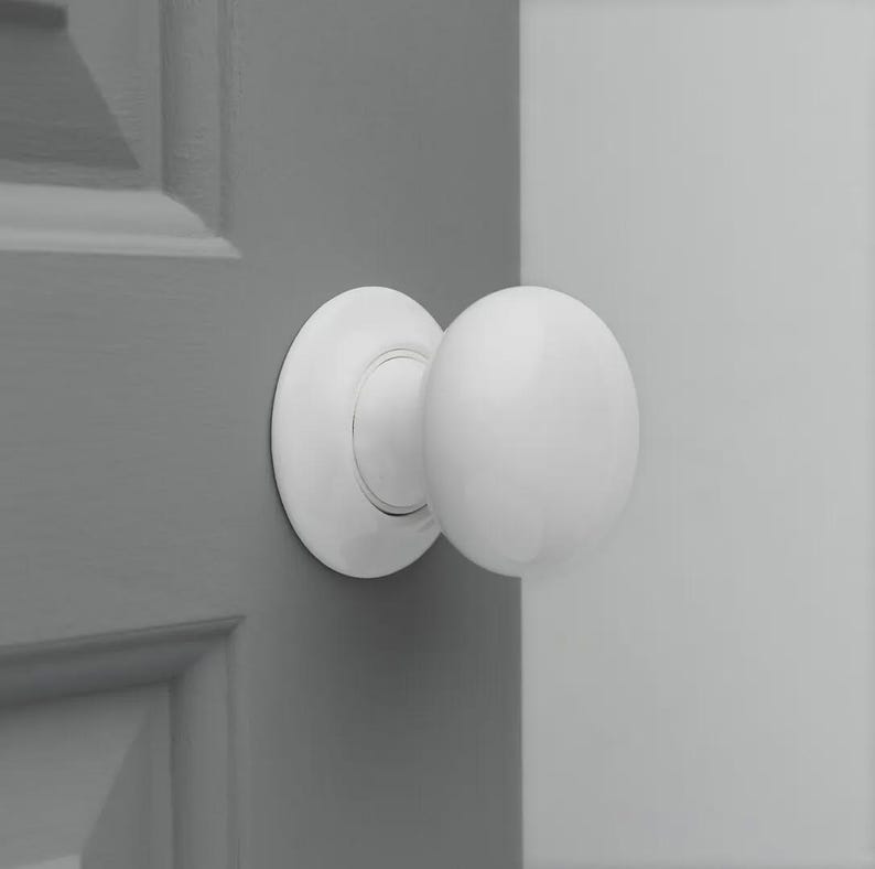 Op de afbeelding: Een witte, ronde deurknop met een bijpassende cirkelvormige achterplaat. De knop is gemonteerd op een grijze deur, met een deel van een witte deur zichtbaar op de achtergrond. De deurknop is een klassiek ontwerp.