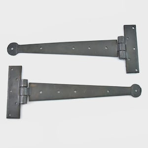 Op de afbeelding: Twee zwarte metalen T-scharnieren. Elk scharnier heeft een rechthoekige plaat met schroefgaten, verbonden met een lange, taps toelopende arm met een afgerond uiteinde. De scharnieren zijn ontworpen voor deuren of poorten.