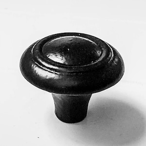 May include: Black cabinet knob with a rounded top and a raised, circular design. The knob has a short, cylindrical base.