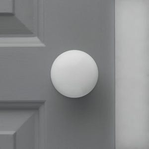 Op de afbeelding: Een witte, bolvormige deurknop is gemonteerd op een grijze deur. De deur heeft een paneelontwerp. De knop is gecentreerd op de deur en lijkt gemaakt van een glad, massief materiaal. De achtergrond is effen grijs.